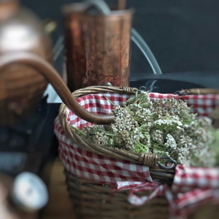 Alambique de cobre junto a una cesta de mimbre con flores de milenrama recién recolectadas.